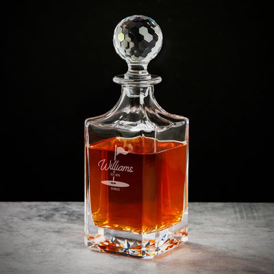 Glass decanter with amber liquid and faceted round stopper, engraved “Williams” and “19 holes,” on gray surface.