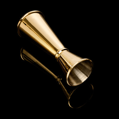 Gold double-sided cocktail jigger lying on a reflective black surface.