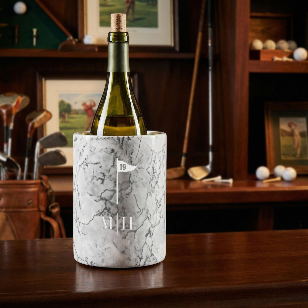 A bottle in a white marble wine chiller engraved with a golf flag icon and initials, set on a wooden table with golf clubs, golf balls, and framed golf artwork in the background